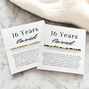 May include: Two white cards with black text that reads "16 Years Loved". Each card has a small bracelet with gold and white beads on one card and gold and black beads on the other card. The text on the cards reads: "For every passing second, minute, day, and year, always remember that you are loved more than words can express. Through every joy and challenge, in every quiet moment and loud celebration, you are surrounded by love and appreciation. No matter where life takes you, you are never alone - your presence makes a difference, and your heart shines bright. So embrace each moment knowing that you are cherished today, tomorrow, and always."