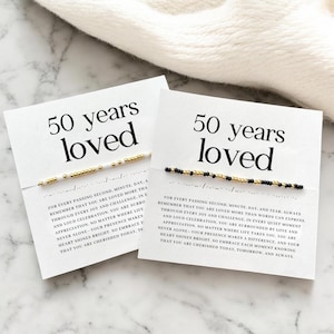 May include: Two white cards with black text that reads "50 years loved". Each card has a beaded bracelet with gold and white beads on one card and gold and black beads on the other card. The text on the cards reads "For every passing second, minute, day, a remember that you are loved more than words can express. Through every joy and challenge, in every quiet moment and loud celebration, you are surrounded by love and appreciation. No matter where life takes you, you are never alone - your presence makes a difference, and your heart shines bright. So embrace each moment knowing that you are cherished today, tomorrow, and always."