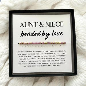 May include: A black box containing a white card with the text "AUNT & NIECE bonded by love". A beaded bracelet with pink and gold beads is displayed. The card also includes a heartfelt message about the bond between an aunt and niece.