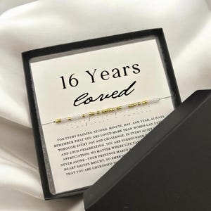 May include: A black gift box containing a card and a bracelet. The card reads "16 Years Loved" with a message below. The bracelet has white and gold beads. The box is open, revealing the contents.