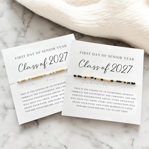 May include: Two white cards with the text "First Day of Senior Year Class of 2027" and a beaded bracelet. One bracelet has white and gold beads, the other has black and gold beads. The cards have inspirational text.