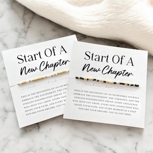 Peut inclure: Deux cartes blanches avec le texte "Start Of A New Chapter" en noir. Chaque carte a un bracelet de perles. Un bracelet a des perles dorées et blanches, l'autre a des perles noires et dorées. Le texte en dessous dit "C'est le début..."
