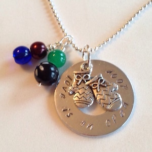 May include: Silver necklace with a charm that says "Love is an open door" with a pair of mittens on it. The necklace has a silver chain and is adorned with blue, green, red, and black beads.