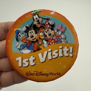 Könnte beinhalten: Ein runder, orangefarbener Button mit dem Text "1st Visit!" und den Worten "Walt Disney World". Der Button zeigt die Zeichentrickfiguren Mickey Mouse, Minnie Mouse, Donald Duck, Goofy, Pluto und Daisy Duck.