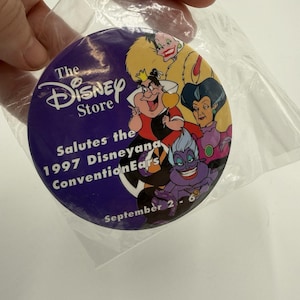 Peut inclure: Un bouton rond violet de Disney Store représentant des méchants Disney. Le bouton indique "The Disney Store Salutes the 1997 Disneyana Convention Ears" et "September 2-6". Le bouton est dans un sac en plastique transparent.