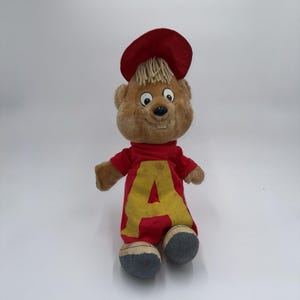May include: A plush toy of a cartoon character with a red shirt featuring a large yellow "A". The character has a brown face, blonde hair, a red hat, and gray and blue shoes. The toy is standing upright.
