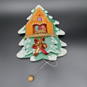 May include: A green wooden Christmas tree ornament with white glitter snow accents. It features an orange house structure in the center with two small figures, a gold ribbon trim, a red bow with two gold bells, and a hanging string with a bead.