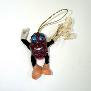 May include: A hanging ornament shaped like a dark purple raisin character with blue eyelids, white gloves, black pipe cleaner limbs, and peach-colored shoes. The figure holds a small cluster of off-white fabric flowers and has a gold hanging cord.