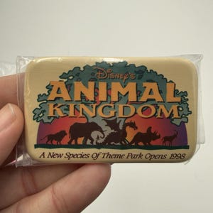 Könnte beinhalten: Ein rechteckiger Ansteck-Pin mit dem Text "Disney's Animal Kingdom" in Orange und Grün, mit Tier-Silhouetten. Der Pin enthält den Text "A New Species Of Theme Park Opens 1998".