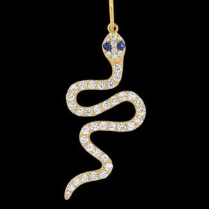 Pode incluir: Um pingente de cobra dourada com um design curvo. O corpo da cobra é incrustado com pequenas pedras preciosas transparentes. Os olhos são duas pequenas pedras preciosas azuis. O pingente pende de um pequeno laço na parte superior.