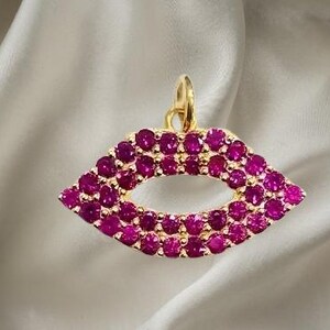 May include: A gold-colored pendant in the shape of lips, encrusted with numerous small, round, magenta-colored gemstones. The pendant hangs from a small loop at the top. The background is a soft, off-white fabric.