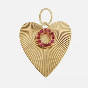 Könnte beinhalten: Goldfarbener Herzanhänger mit strukturiertem, strahlenförmigem Design. Ein Kreis aus kleinen, roten Edelsteinen ist in der Mitte eingesetzt. Der Anhänger hat oben eine Schlaufe für eine Kette. Schmuck.