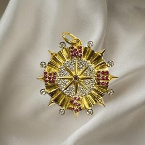 May include: A gold pendant with a starburst design, featuring a central star pattern with clear and red gemstones. The pendant has a loop for a chain and is set against a white, silky background.