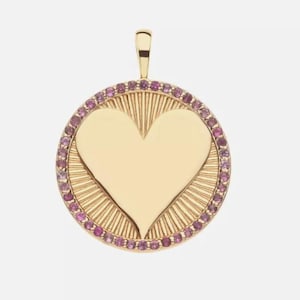 Könnte beinhalten: Goldfarbener Anhänger mit herzförmigem Mittelteil. Das Herz ist von einem Kreis aus kleinen, rosa Edelsteinen umgeben. Der Anhänger hat ein strukturiertes, sonnenstrahlenartiges Design um das Herz und eine kleine Schlaufe.