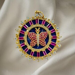 May include: A gold-toned pendant with a butterfly design. The pendant features a blue center with a butterfly made of red gems. The butterfly is surrounded by a circle of small diamonds and pink rectangular gems. The outer edge has small diamond accents.