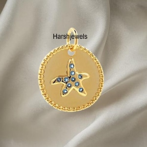 Puede incluir: Colgante dorado con diseño de estrella de mar adornado con piedras preciosas azules. El colgante tiene un borde texturizado y un lazo para una cadena. El texto "Harshjewels" es visible. Forma circular.