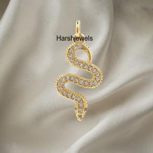 May include: A gold-colored snake pendant with clear, round gemstones. The pendant is in a serpentine shape, with the text "Harshjewels" printed on the background. The pendant is hanging from a small loop at the top.