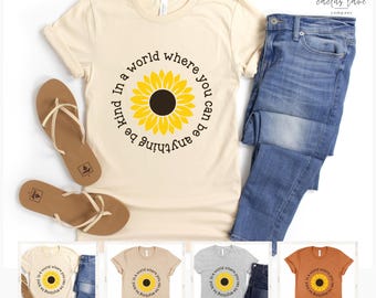 May include: Cream-colored t-shirt with a sunflower design and the text "In a world where you can be anything, be kind." The shirt is paired with blue jeans and brown sandals. Additional shirts in different colors are shown.