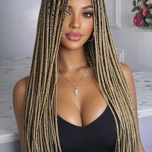 May include: A woman with long, blonde box braids. She wears a black tank top and a gold necklace with a pendant. She has gold hoop earrings and full, pink lips. The background is a light gray.