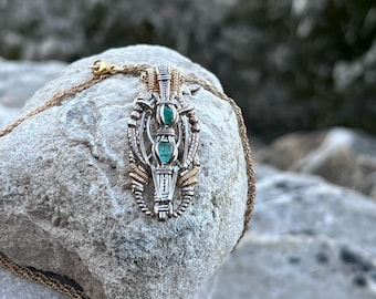 May include: A silver and gold wire wrapped pendant with three green gemstones. The pendant is on a silver chain and resting on a gray rock.