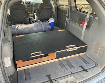 May include: A black wooden storage unit with a natural wood drawer is installed in the back of a minivan. The unit has cut-out handles and is designed to fit the vehicle's cargo area. A tent and cooler are also visible.