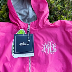 May include: A bright pink children's rain jacket with a hood and a silver zipper. The jacket has a white embroidered monogram and a tag that reads "CHILDRENS NEW ENGLANDER RAIN JACKET". The jacket is from Charles River Apparel.