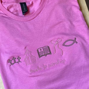 May include: Pink t-shirt with embroidered religious symbols including crosses, a church, an open book, a bow, and a fish. The phrase "Made to worship" is embroidered below the symbols.
