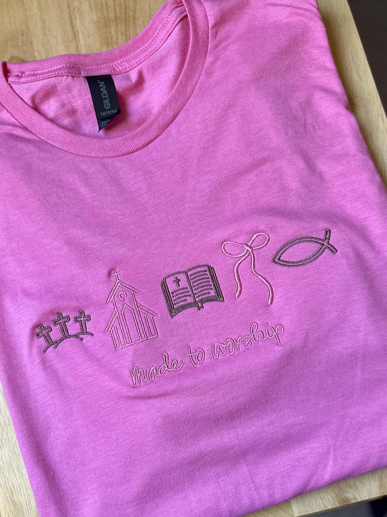 May include: Pink t-shirt with embroidered religious symbols including crosses, a church, an open book, a bow, and a fish. The phrase "Made to worship" is embroidered below the symbols.
