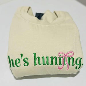 May include: A folded, cream-colored sweatshirt with green embroidered text that reads "he's hunting." A pink bow is embroidered next to the word "hunting."