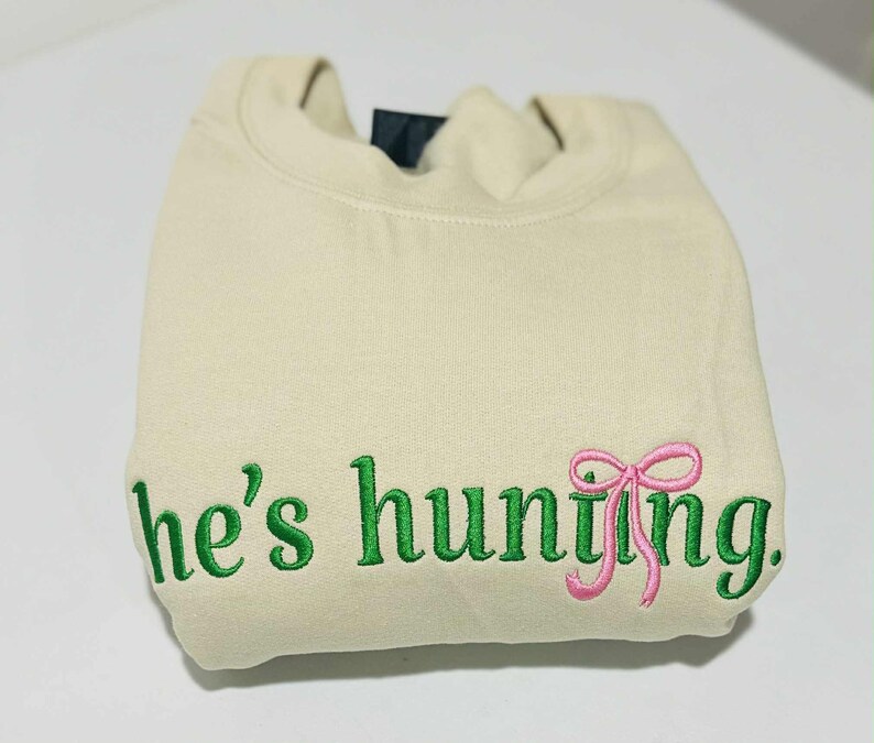 May include: A folded, cream-colored sweatshirt with green embroidered text that reads "he's hunting." A pink bow is embroidered next to the word "hunting."