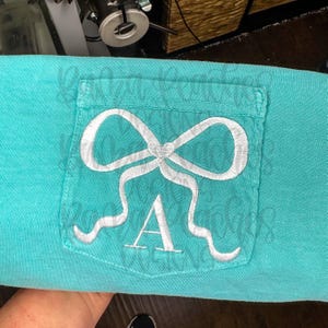 May include: A turquoise shirt with a pocket embroidered with a white bow and the letter "A". The bow design is centered on the pocket, with the letter "A" embroidered below it. The shirt is made of a soft material.