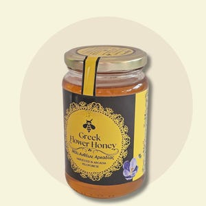 May include: A glass jar of Greek Flower Honey with a gold lid and a black and yellow label. The label features the text "Greek Flower Honey" and "Harvested in Arcadia, Peloponnese", along with a bee illustration and a purple flower.
