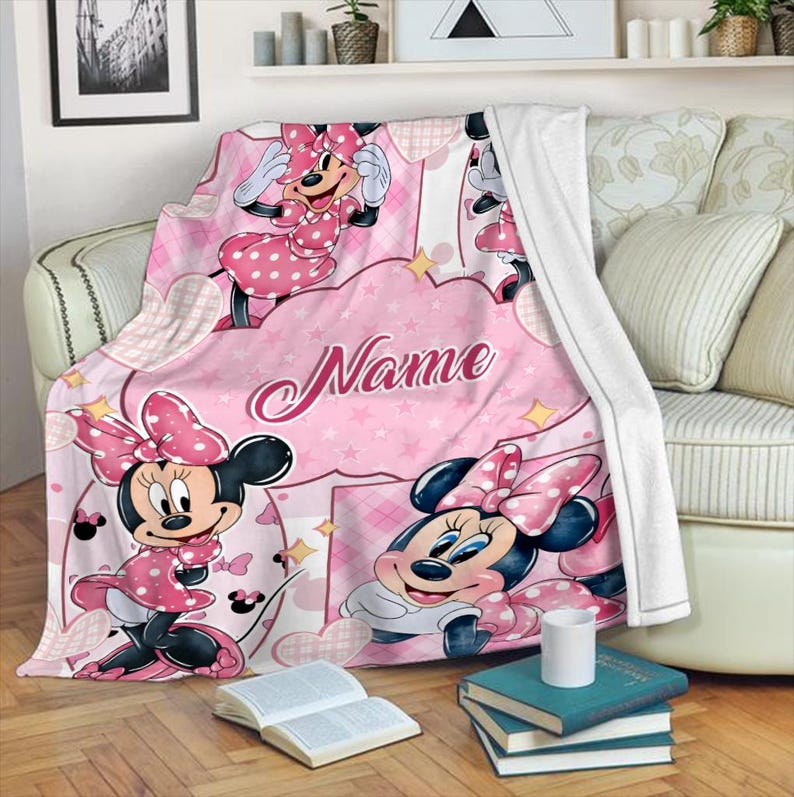 May include: A soft pink fleece blanket featuring Minnie Mouse in various poses, wearing a polka-dot dress and bow. The design includes white heart and star patterns, with the word Name printed in a cursive script across the center of the blanket.