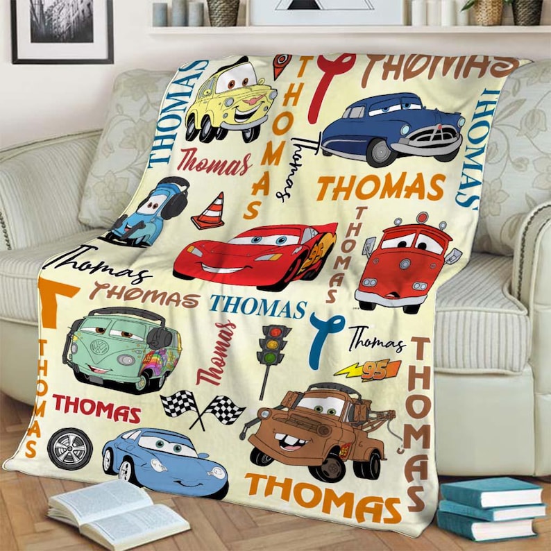 May include: A cream-colored blanket featuring various cartoon car illustrations, including a yellow car, a blue car, and a red race car. The blanket is decorated with the name "Thomas" in multiple places and other car-related graphics.
