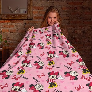 May include: Pink blanket featuring a repeating pattern of Minnie Mouse, bows, and the word "NAME" in black. Minnie Mouse is depicted in her classic red and white polka dot dress and bow. The blanket is held up by a person.