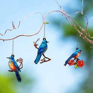 May include: Three hanging stained-glass bird ornaments. Each bird is perched on a branch with flowers. The birds feature blue, orange, and red hues. They hang from a chain and are set against a blurred background of leaves and sky.
