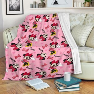 May include: Pink throw blanket featuring a repeating pattern of Minnie Mouse, red polka dot bows, and the word "NAME" in a script font. The blanket is draped over a sofa, with a white backing visible. The design includes white gloves and hearts.