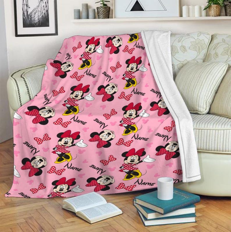 May include: Pink throw blanket featuring a repeating pattern of Minnie Mouse, red polka dot bows, and the word "NAME" in a script font. The blanket is draped over a sofa, with a white backing visible. The design includes white gloves and hearts.
