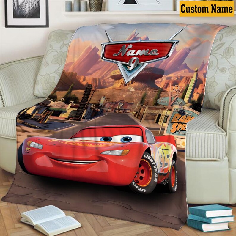 May include: A red Lightning McQueen car blanket with a desert landscape background. The blanket features the text "Name" and the number "9". The blanket is draped over a couch.