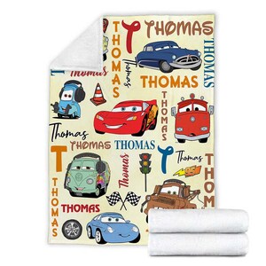 May include: A cream-colored blanket featuring various cartoon cars in red, blue, green, and brown. The name "Thomas" is written in a stylized font throughout the design. The blanket is folded with two white towels.