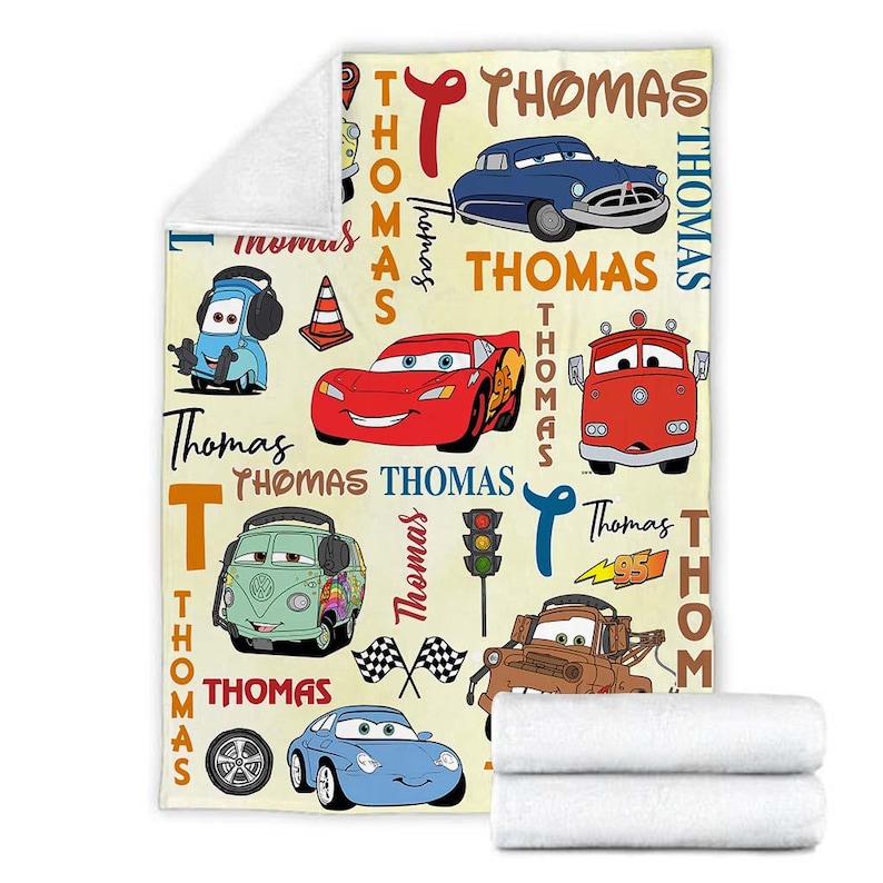May include: A cream-colored blanket featuring various cartoon cars in red, blue, green, and brown. The name "Thomas" is written in a stylized font throughout the design. The blanket is folded with two white towels.