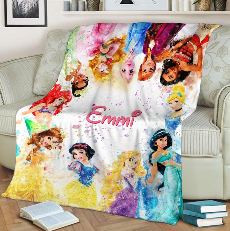 May include: A white blanket featuring watercolor illustrations of various princesses. The blanket has a personalized name, "Emmi," in pink script. The princesses are depicted in their signature outfits, with vibrant colors and details. The blanket is draped over a sofa.