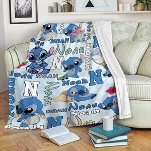 Może przedstawiać: Jasnoniebieski koc z postacią z kresk&oacute;wki Stitch w r&oacute;żnych pozach. Na kocu nadrukowano imię "Noah" i literę "N" w kolorze niebieskim i białym. Koc jest narzucony na kanapę.