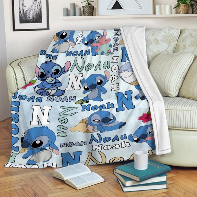 Może przedstawiać: Jasnoniebieski koc z postacią z kresk&oacute;wki Stitch w r&oacute;żnych pozach. Na kocu nadrukowano imię "Noah" i literę "N" w kolorze niebieskim i białym. Koc jest narzucony na kanapę.