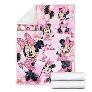 May include: A soft fleece blanket featuring a repeating pattern of Minnie Mouse in a pink polka dot dress, pink bows, butterflies, and balloons against a light pink background. The name Julia is printed in a cursive script in the centre.