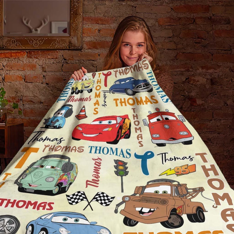 May include: A cream-colored blanket featuring cartoon cars and the name "Thomas" in various fonts. The blanket showcases a red race car, a blue car, and a brown tow truck, among other vehicles. The number "95" is also visible.