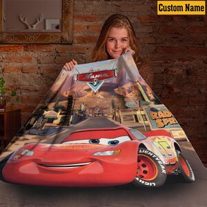 May include: A red Cars-themed blanket featuring Lightning McQueen. The blanket has a detailed image of the character and the Radiator Springs town. The blanket has the text "Name" at the top. The blanket is displayed against a brick wall.