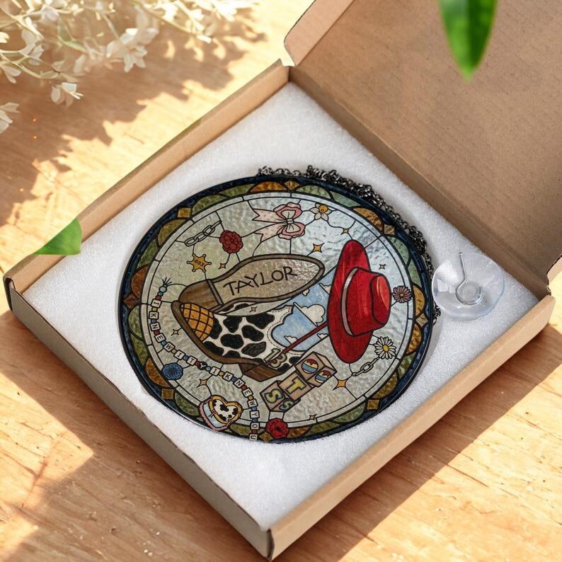 May include: A circular stained glass suncatcher featuring a red hat, a cow-print boot with the name Taylor, and blocks with the letters T, S, and 13. The piece includes a black chain and a clear suction cup, all packaged in a white foam-lined box.