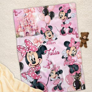May include: A rectangular blanket featuring a light pink background with multiple illustrations of Minnie Mouse, a pink castle, balloons, and butterflies. The name Julia is printed in a cursive script in the centre of the blanket.