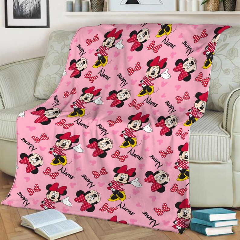 May include: Pink throw blanket featuring a repeating pattern of Minnie Mouse, red polka dot bows, and hearts. The blanket has the word "NAME" printed throughout the design. The blanket is draped over a light-colored sofa.
